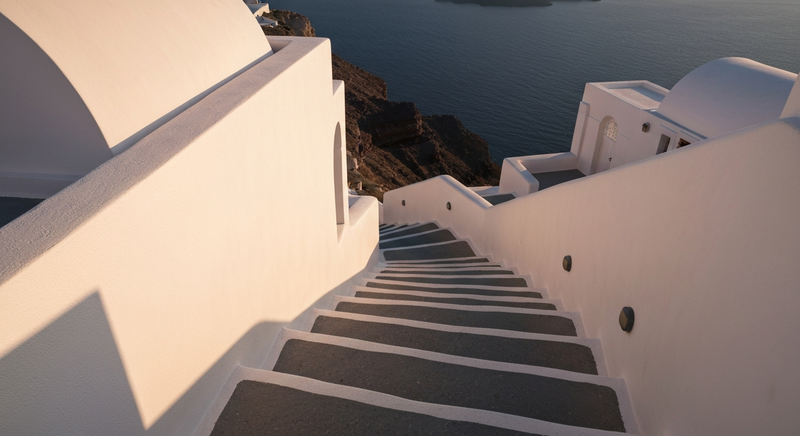 Narrow white stone stairway descending a Santorini cliff at golden hour, caldera visible in the far distance